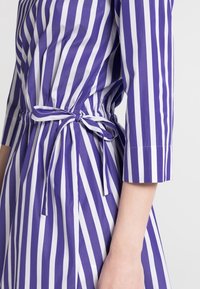 Fialové a bílé pruhované šaty se zúženým pasem a detailním zavazováním. Mají tříčtvrteční rukávy a hladký, lehký materiál.