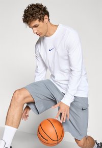 Langærmet hvid Nike skjorte med blåt logo, parret med grå Nike shorts. Modellen holder en basketball, iført hvide sokker og sorte sko.