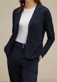 Kvinna klädd i marinblå blazer och matchande byxor, med vit t-shirt med rund halsringning, med händerna i fickorna, mot en enfärgad beige bakgrund.