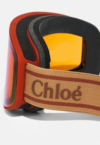 Lunettes de ski rouges avec des verres teintés orange, intérieur en mousse rembourrée et une sangle beige ornée du logo "Chloé" en marron et de rayures rouges.