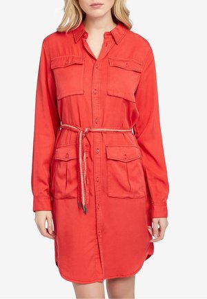 Frau trägt ein rotes Hemdkleid mit langen Ärmeln, vier Klapptaschen vorne und einem dünnen Gürtel, der in der Taille gebunden ist.