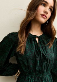 Groene jurk met luipaardprint, een striksluiting bij de hals, een elastische taille en pofmouwen. Zachte stoftextuur en zichtbare gouden accenten.