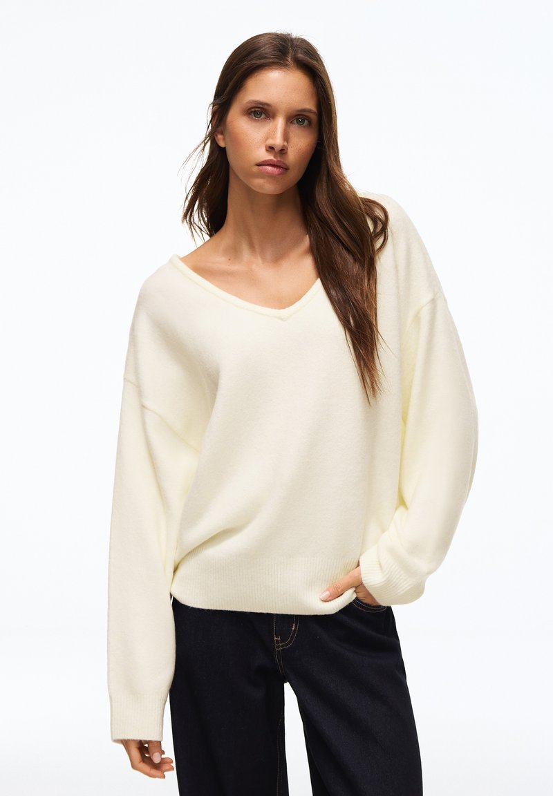 Jeune femme aux longs cheveux bruns portant un pull ample crème à col en V et un pantalon foncé, debout avec une main dans la poche.