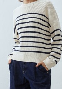 Pull en maille crème avec des rayures horizontales bleu marine, col rond et poignets côtelés. Associé à un pantalon en denim foncé.