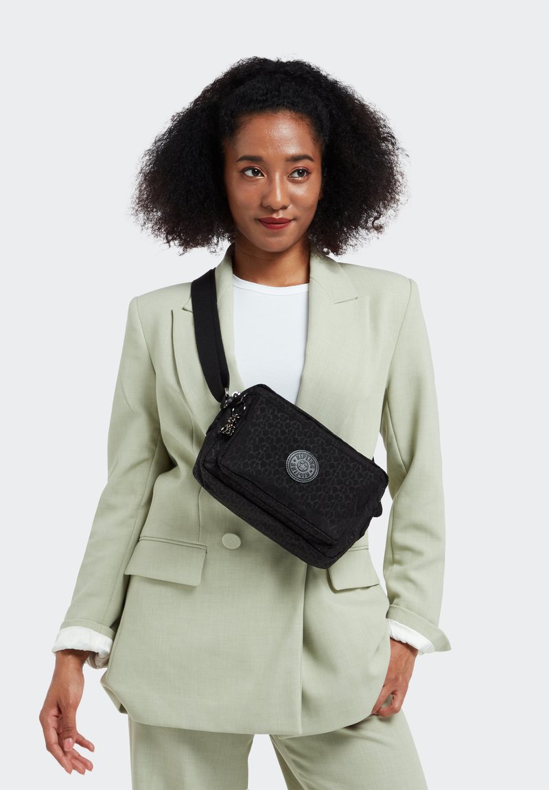 Black rectangular bag with a textured pattern, featuring a round logo, adjustable strap, and zip closure. Worn crossbody over a light green blazer.