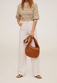 Kobieta w beżowym swetrze dzianinowym i białych szerokich spodniach z wysokim stanem, trzymająca dużą brązową skórzaną torebkę i nosząca sandały z kwadratowym czubkiem.