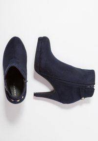 Las botas de tobillo de ante azul marino presentan un cierre de cremallera, punta afilada y un tacón alto y delgado. Están realzadas con pequeños detalles decorativos.