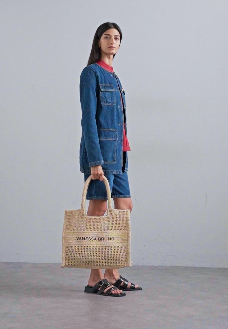 Sac fourre-tout en paille tissée avec le texte noir "VANESSA BRUNO", doté de deux poignées en tissu, associé à une veste en jean bleue et à un short.