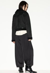 Giacca corta nera con collo in shearling, polsini foderati in pelliccia e cintura in pelle, abbinata a pantaloni neri a gamba larga con pieghe e stivaletti alla caviglia.