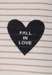 Un tissu rayé en crème et or présente un cœur noir avec le texte "TOMBEZ AMOUREUX" en blanc. Le cœur a une finition texturée.