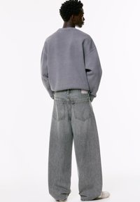 Person von hinten gesehen, der einen übergroßen grauen Pullover, weit geschnittene, ausgebleichte graue Jeans und weiße Sneakers trägt, vor einem weißen Hintergrund stehend.