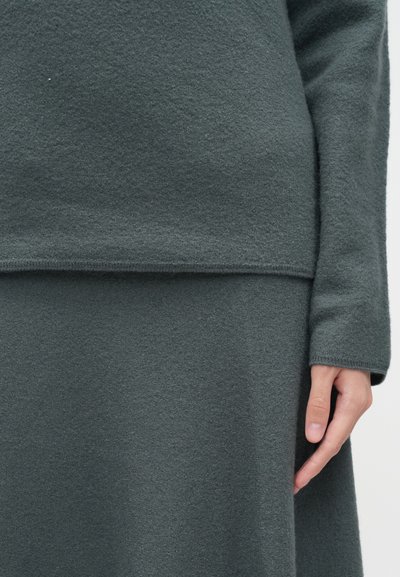 Ένα σκούρο γκρίζο υφασμάτινο ύφασμα σχηματίζει ένα εφαρμοστό τοπ και μια ρέουσα φούστα, με διακριτές ραφές και μακριά μανίκια. Τα χέρια ακουμπούν ελαφρά στο ύφασμα.