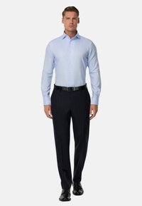 Gai zils pogām apģērbts krekls ar mazu rūtiņu rakstu, kombinēts ar tumši strīpainām biksēm un melniem ādas vīriešu apaviem.