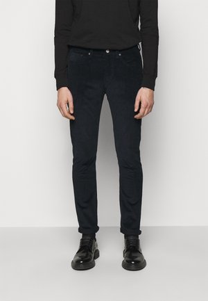 Homme portant un pantalon en velours côtelé noir slim, une chemise noire à manches longues et des chaussures en cuir noires à lacets, debout sur un fond blanc.