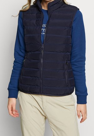 Vrouw die een marineblauw doorgestikt vest draagt over een blauw shirt met lange mouwen en beige broek, staand voor een egale achtergrond.