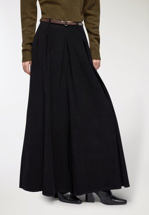 Gonna maxi nera plissettata con una texture liscia, abbinata a una cintura marrone. Indossata con stivaletti neri con tacco, su uno sfondo chiaro.