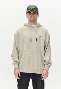 Beige hættebluse med frontlomme og snoretræk. Blødt materiale og løs pasform. Sorte bukser, hænder synlige på siderne.