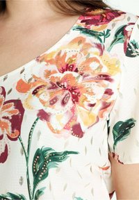 Gros plan sur un tissu blanc à motif floral avec de grandes fleurs orange, rose et verte ornées de petits clous dorés sur un vêtement à manches courtes.