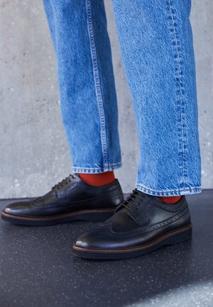 Sorte lædersko med brogue-detaljer, rund tå og snørebånd; parret med blå denimjeans og røde sokker.