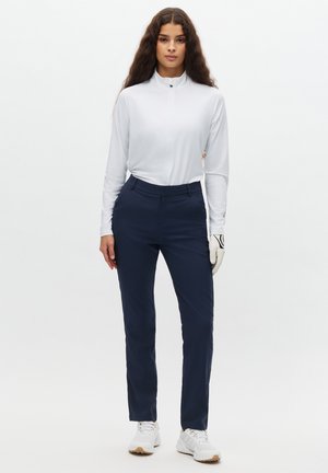 Top bianco a maniche lunghe con collo a zip, pantaloni sartoriali blu navy, guanto da golf bianco e scarpe da ginnastica bianche. Tessuti lisci con un design aderente.