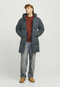 Dark green puffer coat with a hood, featuring a quilted design, zipper pockets, and a relaxed fit, worn over a rust-colored sweater and jeans.
