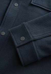 Košile z texturované tkaniny navy modré barvy, se zapínáním na knoflíky na manžetách a černými kovovými patentkami, s tkaným vzorem a přizpůsobeným střihem.