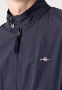 Gros plan d'une personne portant une veste bleu marine avec une fermeture éclair et deux boutons sur le col, avec un petit logo GANT sur la poitrine.