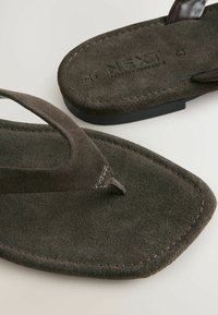 Tamsiai rudos šlepetės su zomšos tekstūra ir išskirtiniu piršto įdėjimu, su prekės ženklu "NEXT" išspausdintu ant pagrindo.