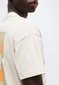 Κοντινό πλάνο ενός ατόμου που φοράει μπλούζα σε μπεζ χρώμα με το κείμενο "TERREX" στο μανίκι και ένα πορτοκαλί, σχεδιαστικό μοτίβο στην πλάτη.