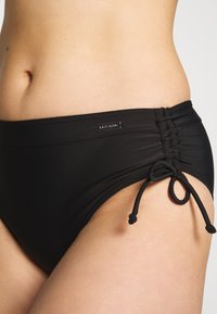 Schwarze Bikinihose aus glattem Material mit gerafftem Seitendetail und verstellbarem Bindeband. Kleine metallene Logo-Plakette ist am Bund sichtbar.