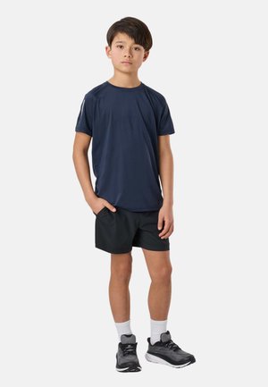 Chłopiec stojący z rękami w kieszeniach, ubrany w granatową sportową koszulkę, czarne spodenki, białe skarpetki i czarne buty do biegania na białym tle.