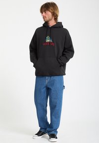 Zwarte hoodie met een kleurrijk grafisch ontwerp op de borst, gecombineerd met een losse spijkerbroeken in blauw en zwarte sneakers.