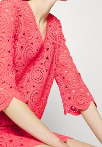 Vestido de ganchillo coral con patrones de encaje en forma circular, escote en V y mangas hasta el codo. Tejido ligero, texturizado con detalles de trama abierta.