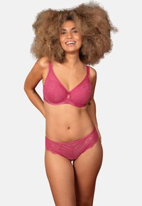 Woman with curly hair smiling, wearing matching pink lace bra and panties, standing against white background with hands behind back.