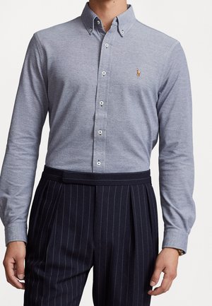 Homme portant une chemise bleu clair à manches longues boutonnée avec un petit logo brodé, rentrée dans un pantalon à rayures foncé taille haute.