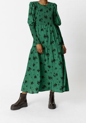 Green floral dress with long puff sleeves and smocked bodice, featuring a flowing tiered skirt, paired with black ankle boots.