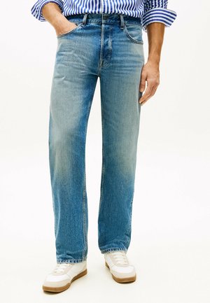 Mann trägt hellblaue Jeans mit geradem Bein, weiße Turnschuhe und ein blau-weiß gestreiftes langärmeliges Hemd, eine Hand in der Tasche.