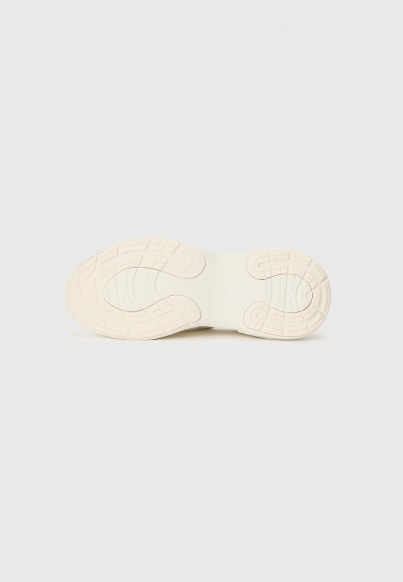 Podeszwa sportowego buta w off-white gumie z teksturowanym wzorem gripu, z zaokrąglonymi sekcjami i liniowymi rowkami zapewniającymi przyczepność.