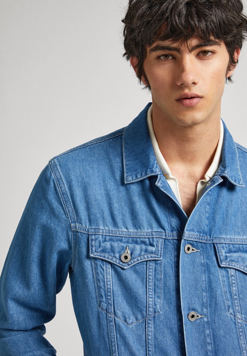 Chaqueta de mezclilla en azul claro con cuello con solapas, cierre de botones y dos bolsillos en el pecho. Presenta una textura suave y un diseño casual.