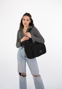 Black messenger bag made of durable fabric, featuring a textured front pocket, adjustable strap, and zip closure. Worn by a model.