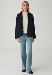 Giacca blu navy in misto lana con colletto, due tasche frontali, indossata sopra un maglione crema chiaro e jeans azzurri a zampa. Calzature nere.