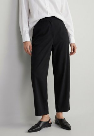 Personne portant un pantalon noir droit, une chemise blanche boutonnée et des chaussures slingback noires pointues, debout devant un fond uni.