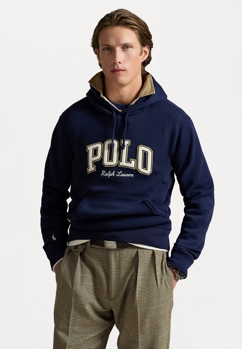 Tengerészeti kapucnis pulóver, "POLO" és "Ralph Lauren" bézs hímzéssel. Kanguru zsebbel és bordázott mandzsettákkal rendelkezik. Kockás nadrággal viselve.