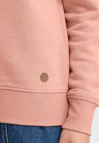 Rosa hettegenser med ribbestrikkede mansjetter og kant, med et brunt lærlogo-patch. Mykt, strukturert stoff og casual passform.