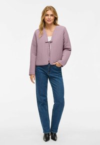 Vadderad lavendeljacka med V-ringning och en framstående rosettdetalj, tillsammans med raka blå jeans och svarta spetsiga stövlar.