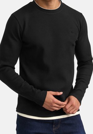 Homme portant un pull côtelé noir à manches longues par-dessus une chemise blanche, les mains jointes devant et un jean bleu visible.