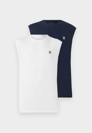 Uomo con maglietta bianca senza maniche, jeans azzurri a gamba larga e sneakers nere, in piedi che guarda indietro oltre la spalla.