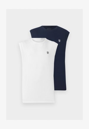 Mann med hvit ermeløs skjorte, lyseblå vide jeans og svarte joggesko, stående og ser tilbake over skulderen.