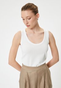 Top de tirantes blanco con textura acanalada y escote redondeado, llevado con una falda plisada de cintura alta de color marrón claro. Textura de tela suave.