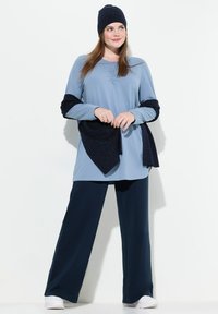 Tunika azzurra chiara con maniche lunghe e dettagli blu navy, abbinata a pantaloni larghi blu scuro e un berretto lavorato a maglia coordinato. Tessuto di consistenza liscia.